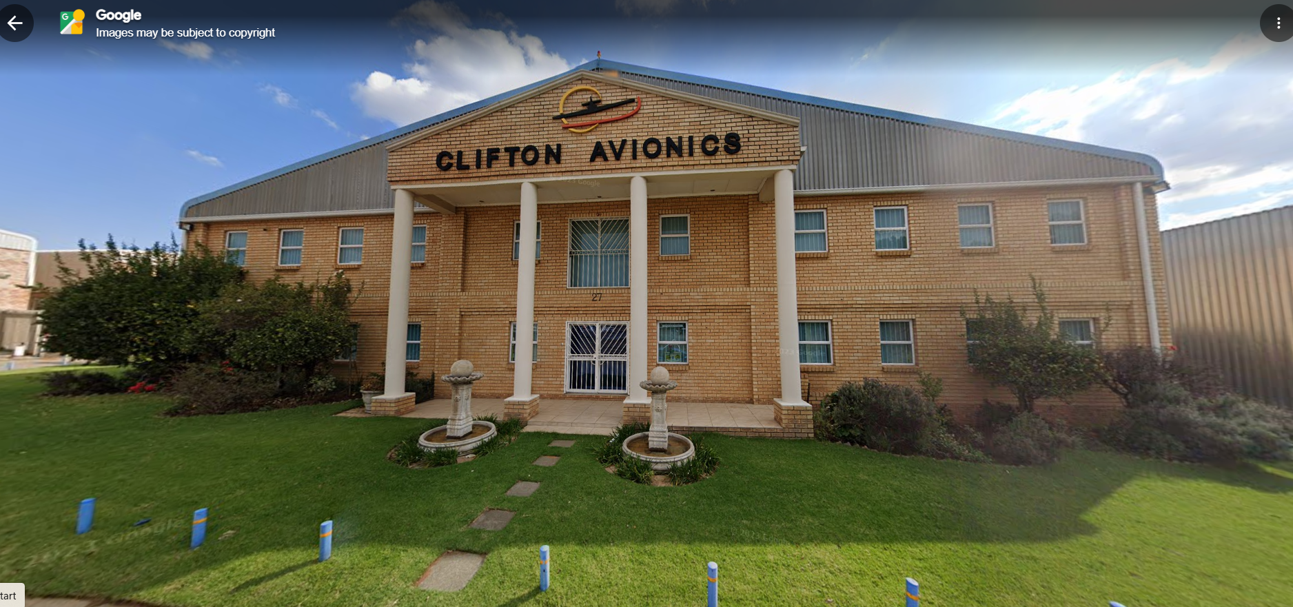 Image shows a large, European style, facebrick building attached to an airport hangar with a lush green garden out front