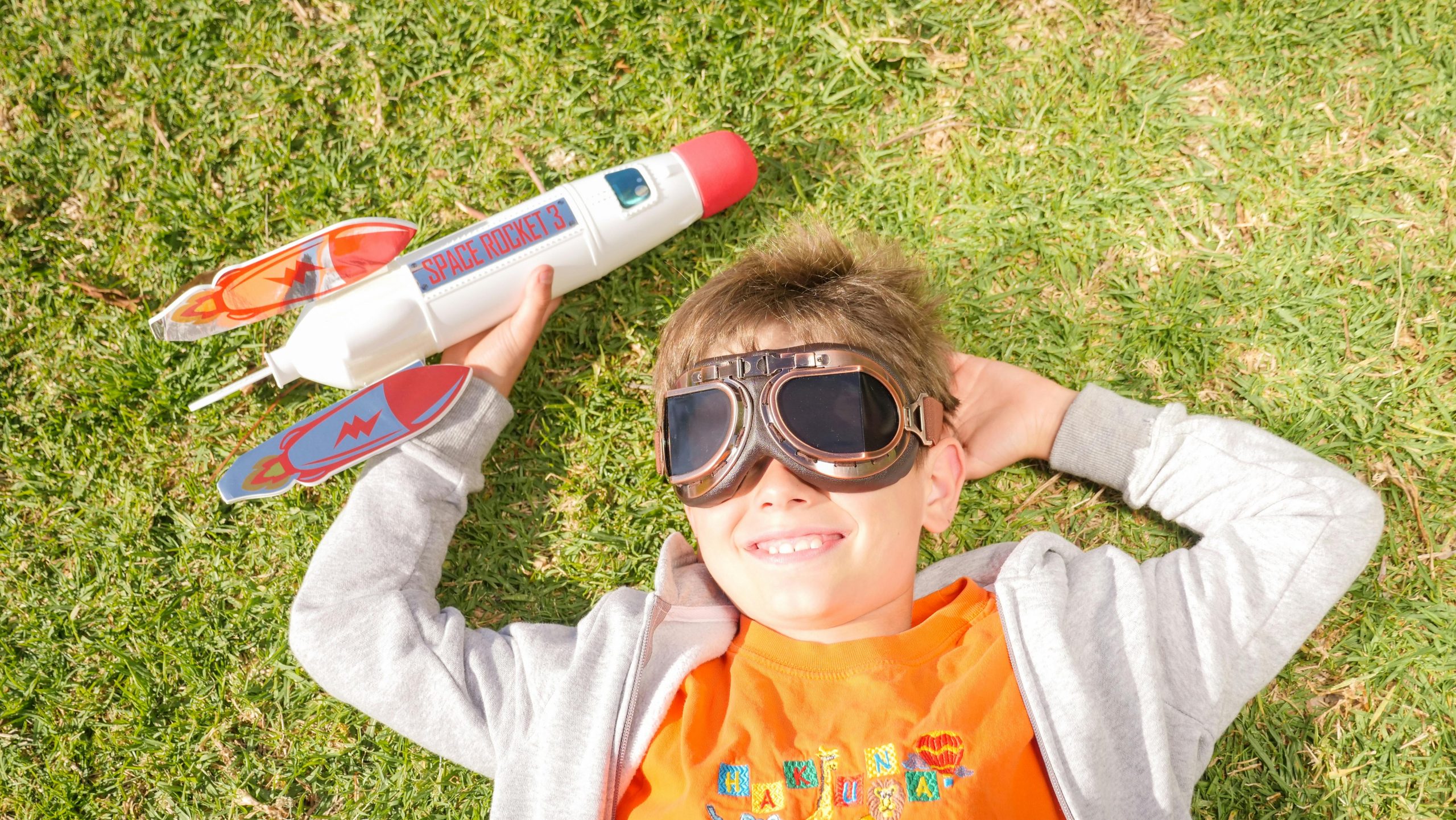 A young boy lying on grass, wearing goggles, and playing with a toy rocket, smiling in the sun.